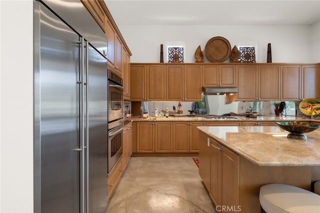 56102 Baltusrol Ln, Quinta, CA 92253 - kitchen with appliances with stainless steel finishes, finished concrete flooring, light stone countertops, under cabinet range hood, and a breakfast bar