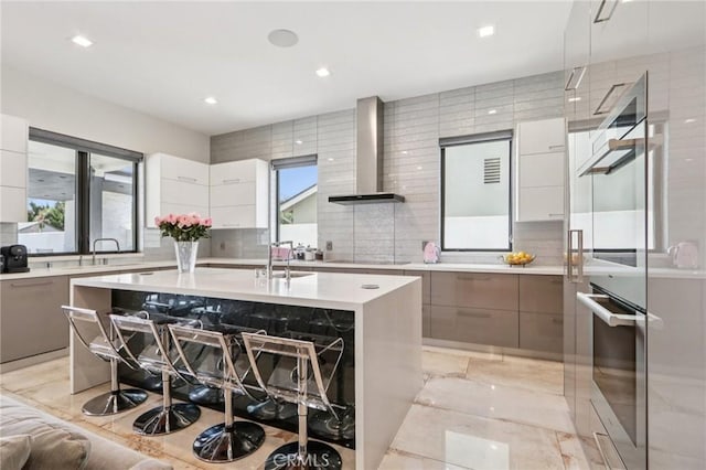 10418 Rubio Ave, Granada Hills, CA 91344 - kitchen featuring modern cabinets, wall chimney exhaust hood, light countertops, a kitchen island with sink, and stainless steel double oven