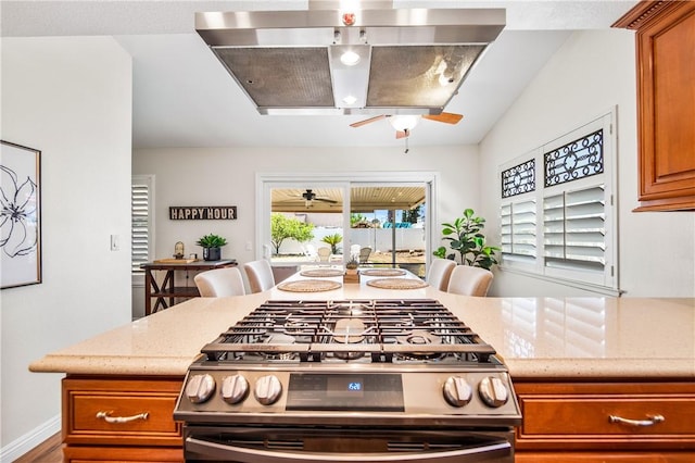 8761 Sandhill Dr, Riverside, CA 92508 - kitchen featuring stainless steel range with gas cooktop, ceiling fan, brown cabinets, wall chimney range hood, and light stone countertops