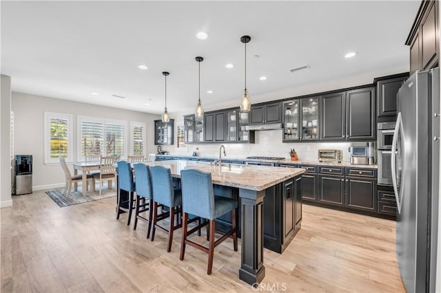 33303 Barmetta Ln, Temecula, CA 92592 - kitchen featuring appliances with stainless steel finishes, a center island with sink, light wood finished floors, under cabinet range hood, and light stone counters