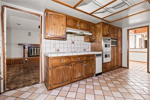 42865 Normandy Ln, Lancaster, CA 93536 - kitchen with white double oven, backsplash, a brick fireplace, brown cabinets, and under cabinet range hood