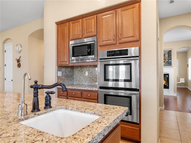7219 Evening Star Rose Ct, Oak Hills, CA 92344 - kitchen with appliances with stainless steel finishes, arched walkways, light stone counters, tasteful backsplash, and brown cabinetry