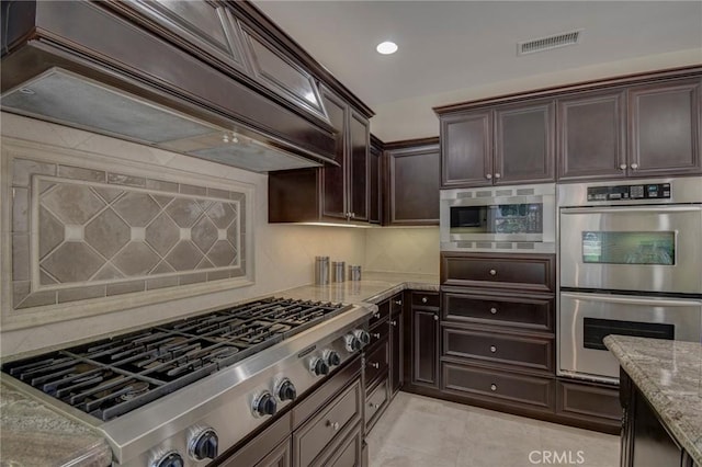 62 Gentry, Irvine, CA 92620 - kitchen featuring appliances with stainless steel finishes, premium range hood, light stone counters, dark brown cabinetry, and backsplash