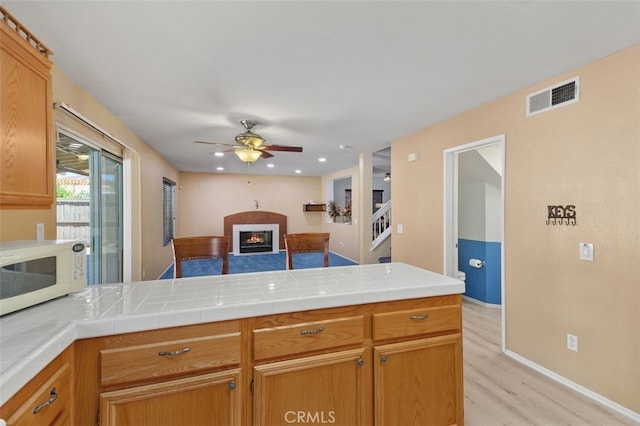 23050 Catt Rd, Wildomar, CA 92595 - kitchen featuring white microwave, light wood-style flooring, a peninsula, a glass covered fireplace, and ceiling fan