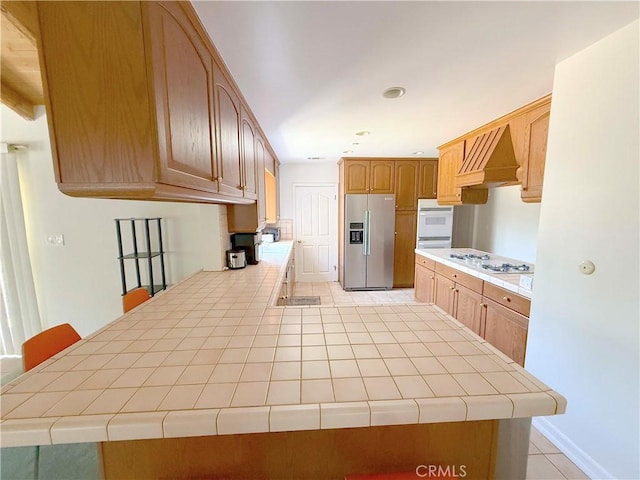 21045 Hemmingway St, Canoga Park, CA 91304 - kitchen with white appliances, premium range hood, a peninsula, light tile patterned floors, and tile counters