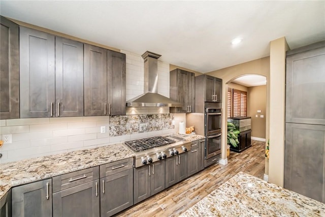 29155 Celestial Dr, Menifee, CA 92584 - kitchen featuring wall chimney range hood, appliances with stainless steel finishes, arched walkways, light wood-type flooring, and tasteful backsplash