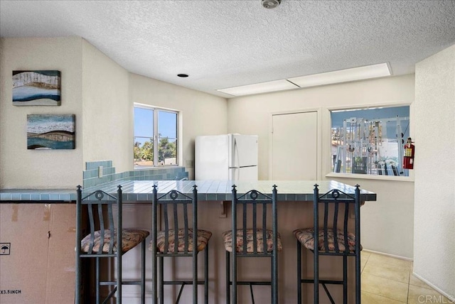 7394 Altiva Pl, Carlsbad, CA 92009 - kitchen featuring freestanding refrigerator, tile counters, a breakfast bar, light tile patterned flooring, and a textured ceiling