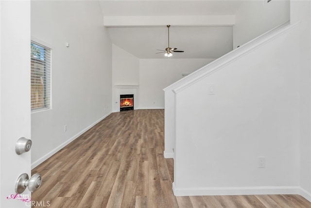 empty room with ceiling fan, wood finished floors, a warm lit fireplace, high vaulted ceiling, and beam ceiling