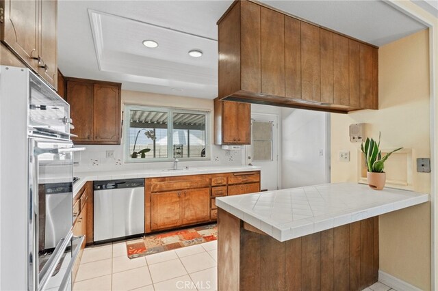 3186 N Hartman St, Orange, CA 92865 - kitchen featuring appliances with stainless steel finishes, a tray ceiling, brown cabinetry, a peninsula, and tile countertops