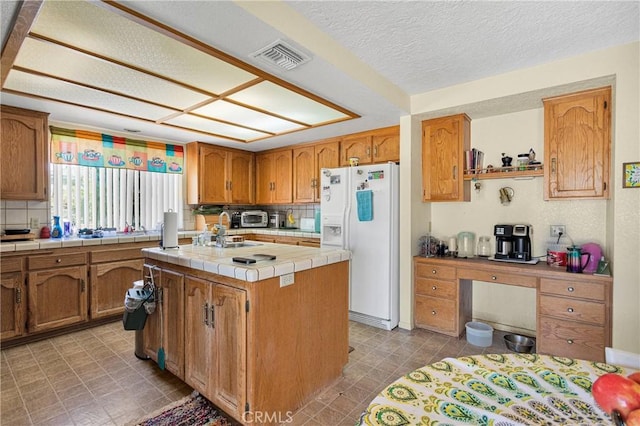 19428 Shasta Rd, Apple Valley, CA 92307 - kitchen with white refrigerator with ice dispenser, a kitchen island with sink, tile countertops, brown cabinetry, and a textured ceiling