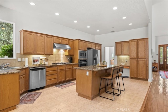 12703 Paso Verde Ct, Atascadero, CA 93422 - kitchen with appliances with stainless steel finishes, under cabinet range hood, dark stone counters, a kitchen island, and brown cabinetry