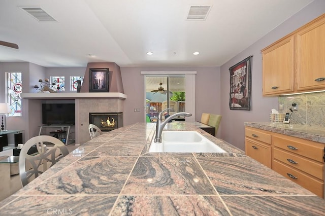 11180 Bridgefield Ct, Riverside, CA 92505 - kitchen with light brown cabinetry, ceiling fan, a warm lit fireplace, backsplash, and healthy amount of natural light