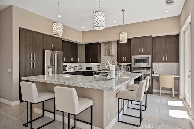 56720 Jack Nicklaus Blvd, La Quinta, CA 92253 - kitchen featuring stainless steel appliances, wall chimney range hood, a kitchen breakfast bar, dark brown cabinetry, and decorative backsplash