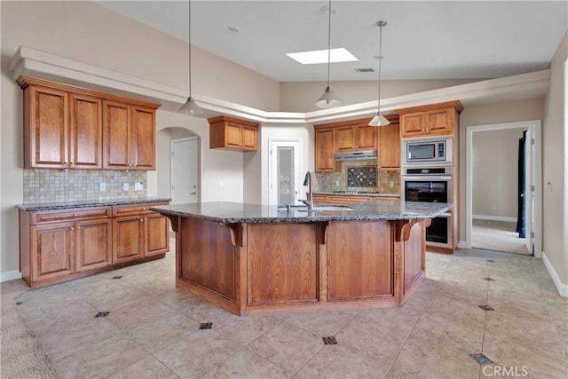 15090 Olema Rd, Apple Valley, CA 92307 - kitchen featuring under cabinet range hood, brown cabinets, appliances with stainless steel finishes, decorative backsplash, and dark stone counters