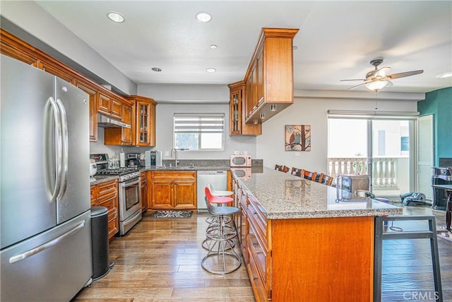 1026 S Oxford Ave Unit 206, Los Angeles, CA 90006 - kitchen with stainless steel appliances, brown cabinetry, a kitchen breakfast bar, ceiling fan, and light stone counters