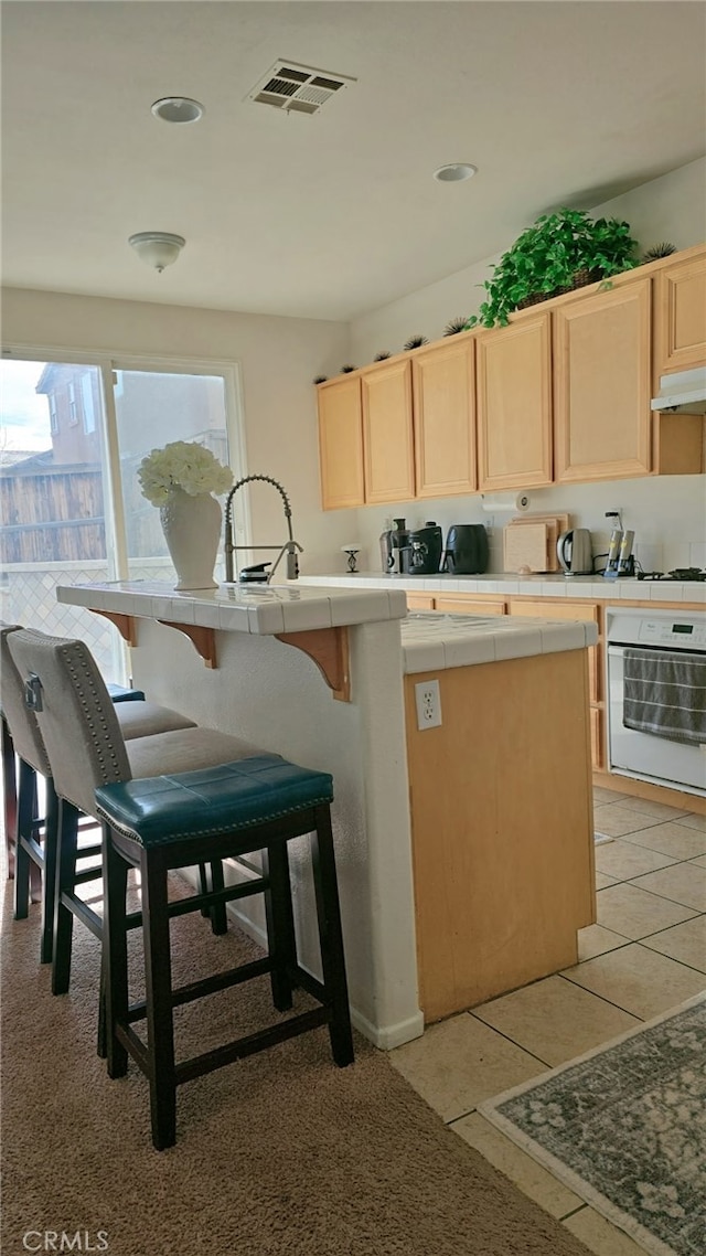 13749 Starshine Dr, Victorville, CA 92392 - kitchen featuring oven, tile countertops, light brown cabinetry, under cabinet range hood, and a breakfast bar