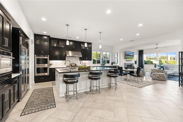 41783 Valencia Way, Temecula, CA 92592 - kitchen featuring wall chimney range hood, appliances with stainless steel finishes, a fireplace, open floor plan, and a breakfast bar area