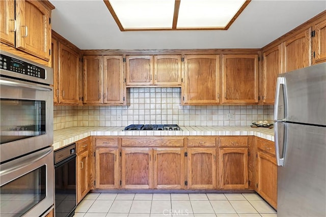 524 St Kevin Ct, Merced, CA 95348 - kitchen with stainless steel appliances, tile countertops, tasteful backsplash, light tile patterned flooring, and brown cabinets