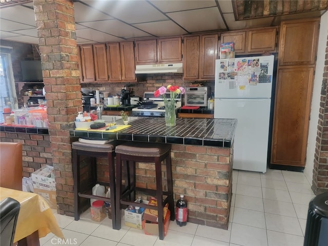 8502 Borson St, Downey, CA 90242 - kitchen featuring freestanding refrigerator, tile counters, under cabinet range hood, light tile patterned flooring, and brown cabinetry