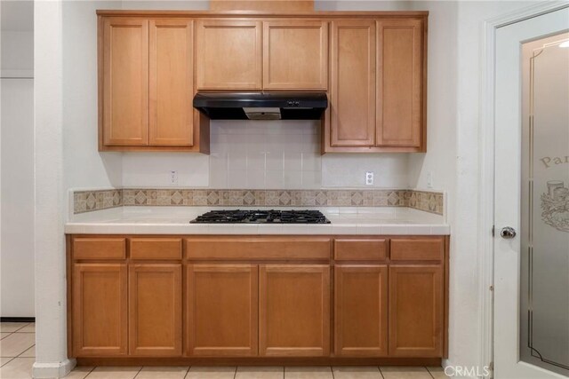 6631 Aggies Ct, Winton, CA 95388 - kitchen with under cabinet range hood, stainless steel gas cooktop, tile counters, brown cabinets, and light tile patterned flooring