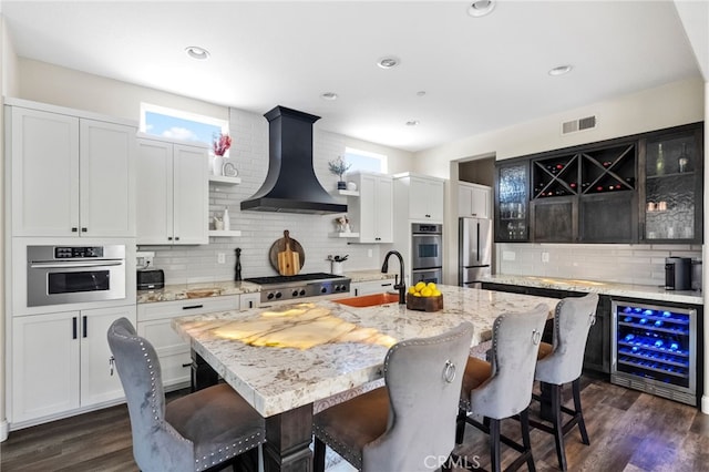 982 Camino Del Arroyo Dr Unit 49, San Marcos, CA 92078 - kitchen featuring wall chimney exhaust hood, wine cooler, decorative backsplash, an island with sink, and dark wood-style flooring