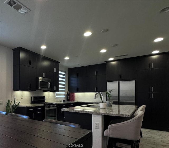 58 Origin, Irvine, CA 92618 - kitchen featuring dark cabinets, stainless steel appliances, tasteful backsplash, an island with sink, and recessed lighting