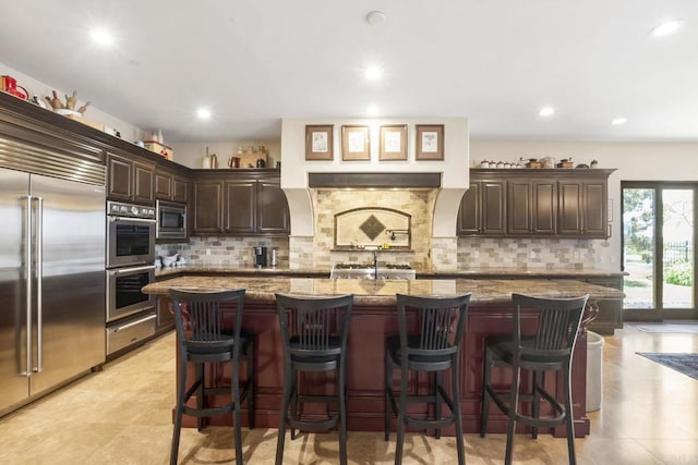 14786 Cool Valley Ranch Rd, Valley Center, CA 92082 - kitchen featuring built in appliances, dark brown cabinetry, tasteful backsplash, an island with sink, and a kitchen bar