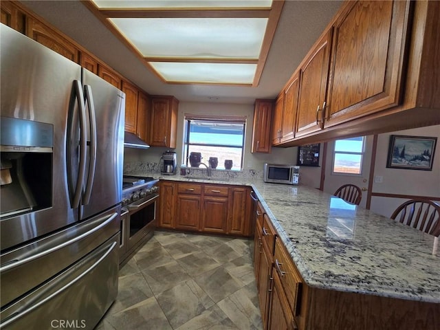 7050 Ivanpah Ave Unit 29, Palms, CA 92277 - kitchen featuring stainless steel appliances, brown cabinetry, a peninsula, light stone countertops, and under cabinet range hood