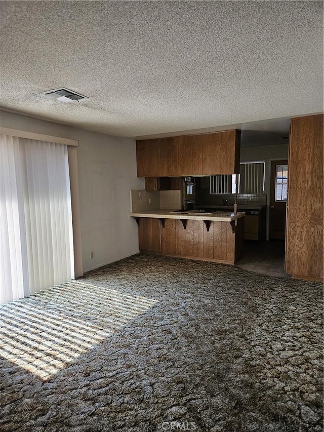 12546 Rain Shadow Rd, Victorville, CA 92395 - kitchen with a peninsula, carpet, a kitchen breakfast bar, a textured ceiling, and brown cabinets