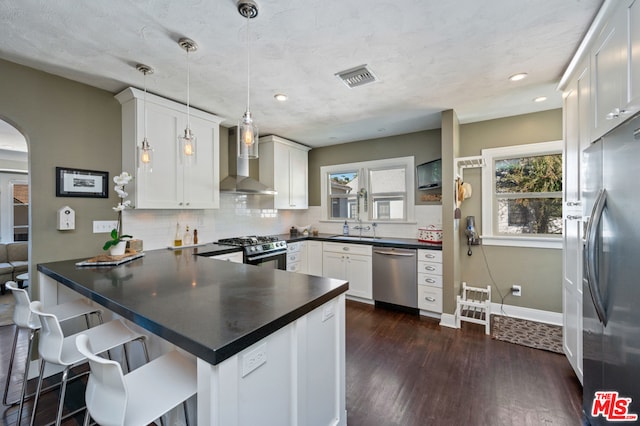 kitchen with kitchen peninsula, wall chimney exhaust hood, appliances with stainless steel finishes, hanging light fixtures, and dark hardwood / wood-style floors