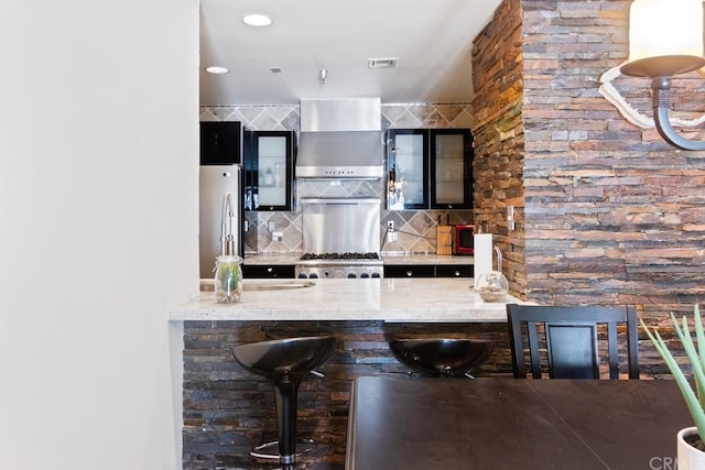 kitchen featuring light stone countertops, appliances with stainless steel finishes, a kitchen breakfast bar, tasteful backsplash, and wall chimney range hood