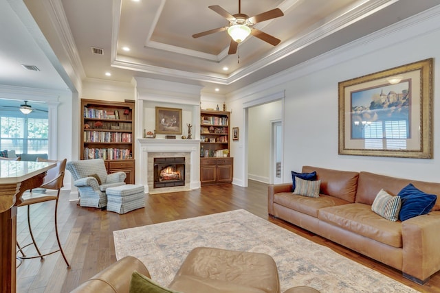 living room featuring hardwood / wood-style flooring, a tiled fireplace, ornamental molding, ceiling fan, and a tray ceiling