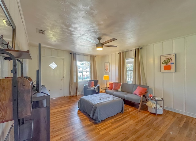 living area with ceiling fan, light wood finished floors, visible vents, and a decorative wall