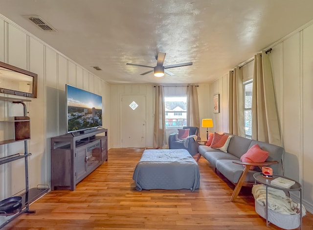 living area with light wood-style floors, visible vents, ceiling fan, and a decorative wall