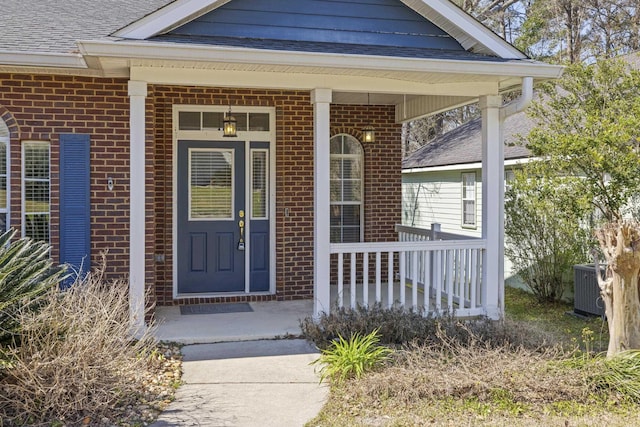 doorway to property with covered porch, brick siding, a shingled roof, and cooling unit