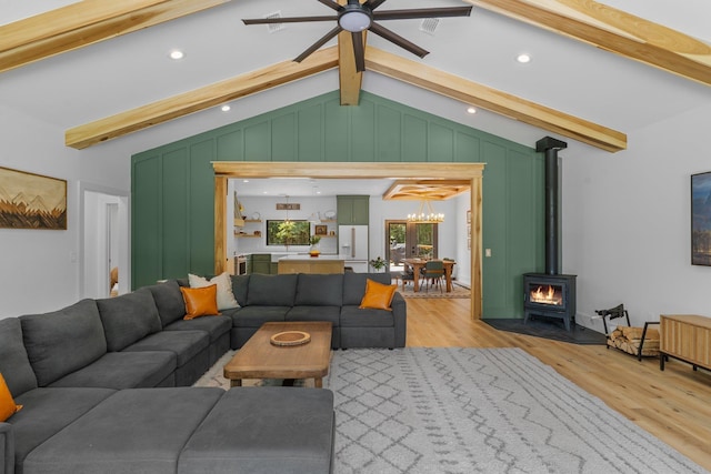 living room featuring ceiling fan with notable chandelier, a wood stove, light hardwood / wood-style flooring, and vaulted ceiling with beams