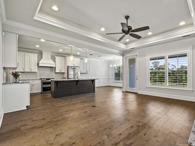 kitchen with appliances with stainless steel finishes, premium range hood, a raised ceiling, a kitchen island with sink, and white cabinets