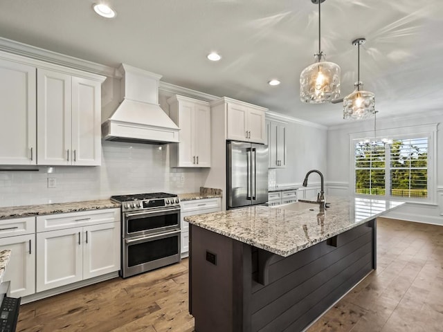 kitchen featuring premium range hood, a kitchen island with sink, white cabinets, decorative light fixtures, and stainless steel appliances