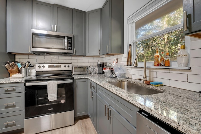 kitchen featuring gray cabinetry, a sink, appliances with stainless steel finishes, backsplash, and light stone countertops