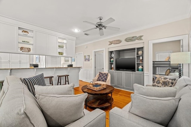 living room featuring built in shelves, light hardwood / wood-style flooring, ceiling fan, and crown molding