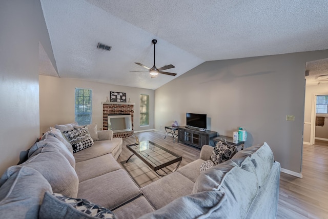 living room featuring ceiling fan, a brick fireplace, light wood-type flooring, lofted ceiling, and a textured ceiling