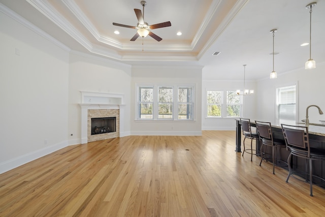 living room featuring a stone fireplace, crown molding, light hardwood / wood-style flooring, a tray ceiling, and ceiling fan with notable chandelier