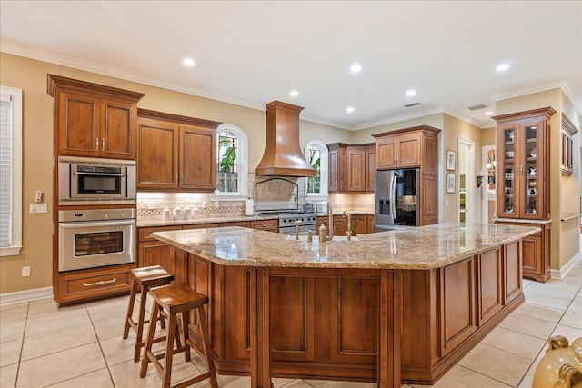 kitchen featuring appliances with stainless steel finishes, tasteful backsplash, light stone countertops, custom exhaust hood, and a large island with sink