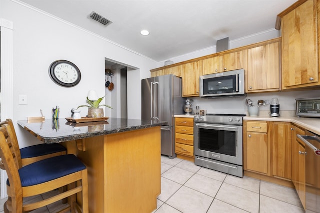 kitchen with light tile patterned floors, appliances with stainless steel finishes, dark stone counters, ornamental molding, and a breakfast bar