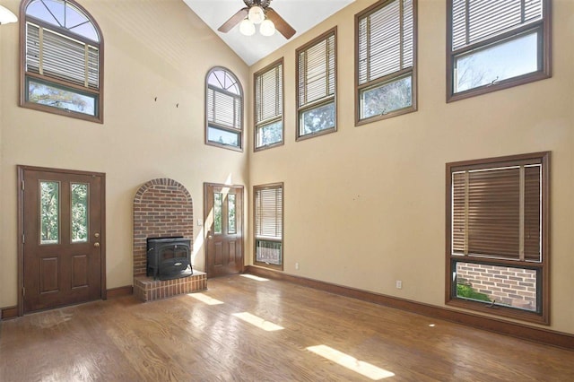 unfurnished living room with high vaulted ceiling, baseboards, wood finished floors, a ceiling fan, and a wood stove