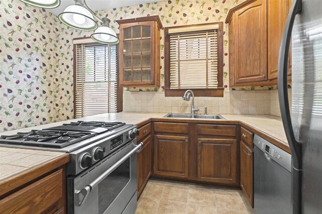 kitchen featuring stainless steel appliances, wallpapered walls, a sink, tile countertops, and glass insert cabinets