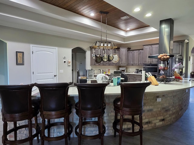 kitchen featuring arched walkways, a tray ceiling, island exhaust hood, a breakfast bar area, and light countertops