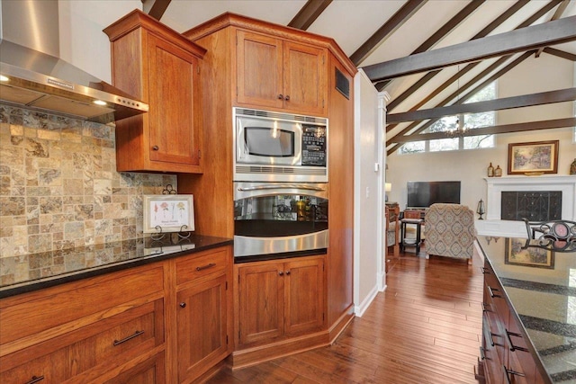 kitchen with lofted ceiling with beams, brown cabinets, appliances with stainless steel finishes, and wall chimney range hood