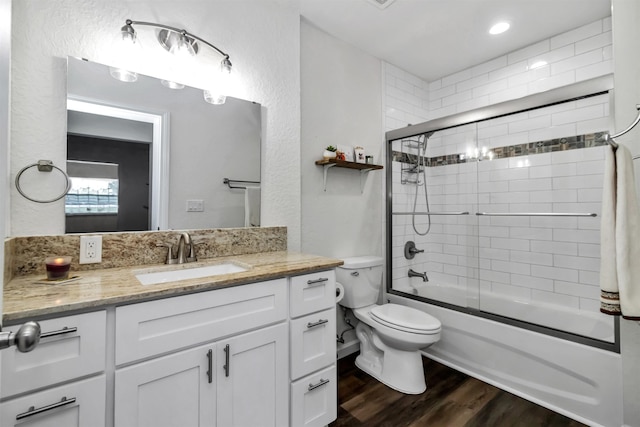 full bath with a textured wall, toilet, shower / bath combination with glass door, wood finished floors, and vanity