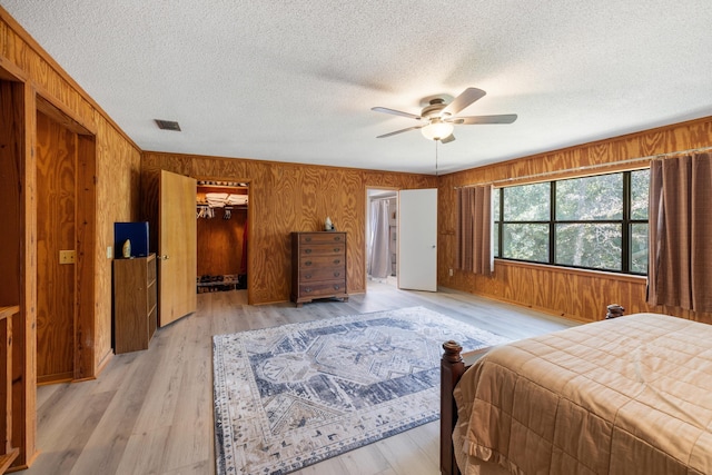 bedroom with light wood-style flooring, a textured ceiling, wood walls, ceiling fan, and a walk in closet